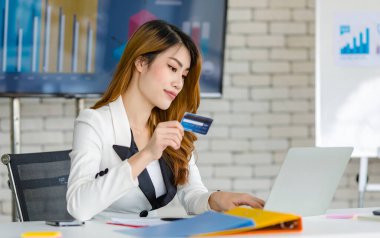 Asyalı, genç, mutlu, güzel, bin yıllık profesyonel başarılı, takım elbiseli kadın iş kadını elinde dizüstü bilgisayarla elinde kredi kartıyla oturmuş gülümsüyor..