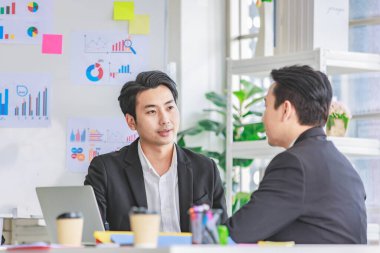 İki Asyalı profesyonel iş adamı resmi takım elbiseli, şirket müşteri brifinglerini dinlerken dizüstü bilgisayar kullanarak toplantı odasında birlikte oturuyorlar..