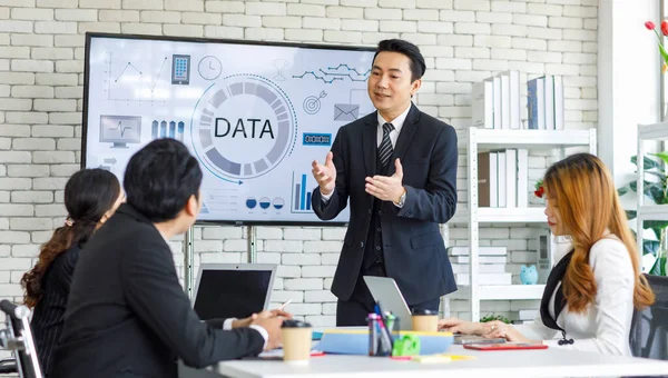 Asian millennial professional successful businessman in black formal suit standing presenting showing company data on big monitor screen with male and female colleagues in meeting room workstation.