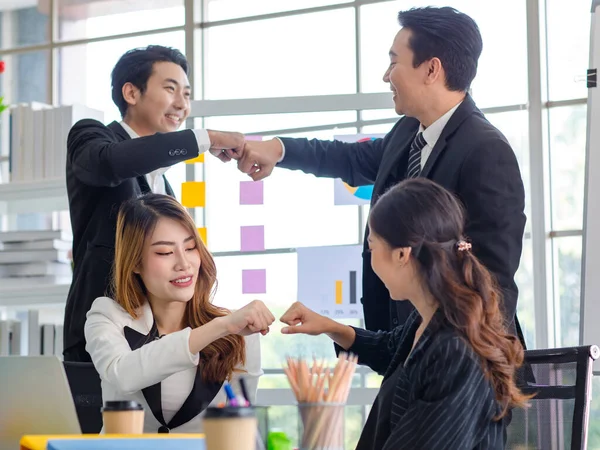 Group of happy Asian millennial professional successful male businessman and female businesswoman in formal suit sitting standing together holding hands fists bumping when working deal agreement done.