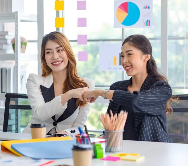 Two happy cheerful Asian millennial professional successful female businesswomen in formal suit sitting together holding hands fists bumping celebrating job agreement deal done at office workstation.