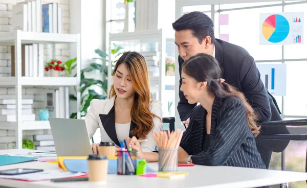 Asian millennial professional successful male businessman mentor in black formal suit standing helping two female employee sitting working with laptop computer at workstation in company meeting room.