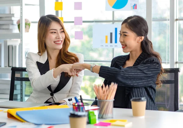 Two happy cheerful Asian millennial professional successful female businesswomen in formal suit sitting together holding hands fists bumping celebrating job agreement deal done at office workstation.