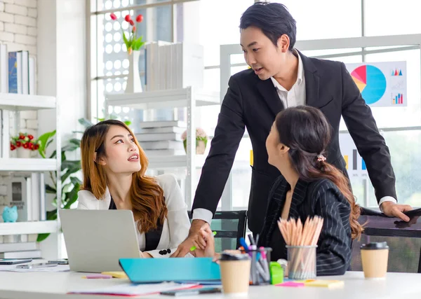 Asian millennial professional successful male businessman mentor in black formal suit standing helping two female employee sitting working with laptop computer at workstation in company meeting room.