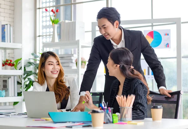 Asian millennial professional successful male businessman mentor in black formal suit standing helping two female employee sitting working with laptop computer at workstation in company meeting room.