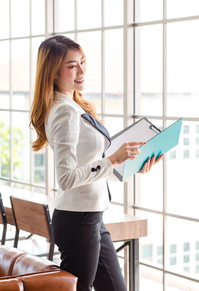 Asian young happy cheerful beautiful millennial professional successful female businesswoman secretary in formal suit stand smiling holding document folder at working counter with blank screen laptop.