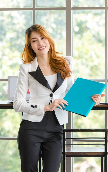 Asian young happy cheerful beautiful millennial professional successful female businesswoman secretary in formal suit stand smiling holding document folder at working counter with blank screen laptop.