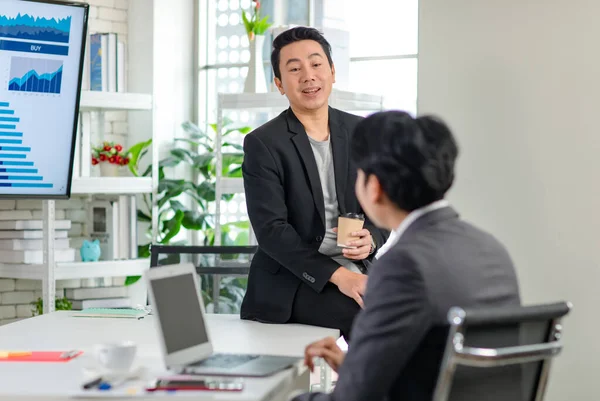 Asian millennial professional successful male businessman in casual suit sitting on table take break drinking coffee talking discussing with unrecognizable unknown colleague in office meeting room.