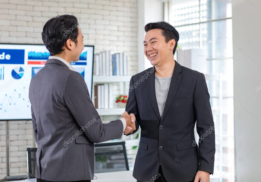 Asian happy cheerful handsome millennial professional successful male businessman in casual blazer standing smiling handshaking greeting with colleague in formal suit in company office meeting room.