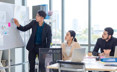 Businessman manager presenter in formal suit standing holding pen pointing at graph chart document on whiteboard presenting company information to Asian male female colleagues in meeting room.