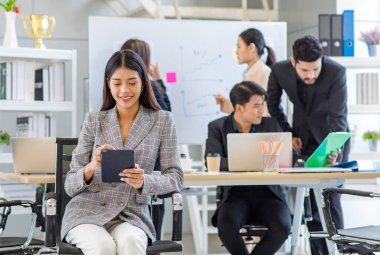 Portrait shot Asian young successful friendly businesswoman in formal suit sitting smiling holding tablet while male businessman and female colleagues working brainstorming together in meeting room.