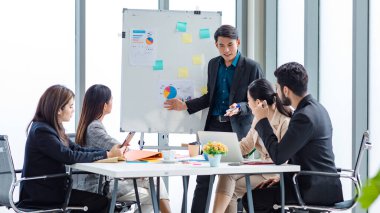 Businessman manager presenter in formal suit standing holding pen pointing at graph chart document on whiteboard presenting company information to Asian male female colleagues in meeting room.
