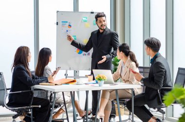 Indian businessman manager presenter in formal suit standing holding pen pointing at graph chart document on whiteboard presenting company information to Asian male female colleagues in meeting room.
