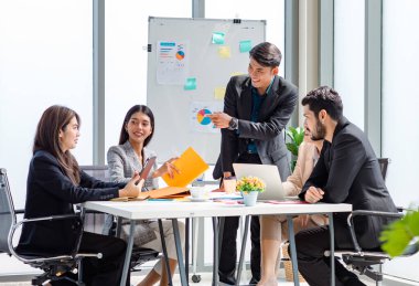 Businessman manager presenter in formal suit standing holding pen pointing at graph chart document on whiteboard presenting company information to Asian male female colleagues in meeting room.