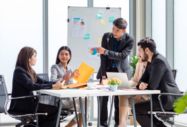 Businessman manager presenter in formal suit standing holding pen pointing at graph chart document on whiteboard presenting company information to Asian male female colleagues in meeting room.