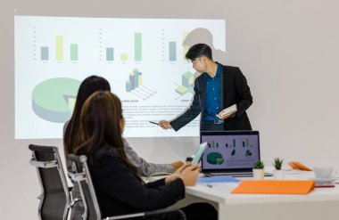 Millennial Asian young cheerful male businessman presenter in formal suit standing holding notebook and pen pointing presenting corporate graph chart data information from wall screen in meeting room.