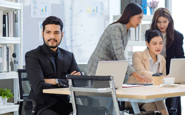 Millennial Asian Indian male confident businessman in formal suit sitting crossed arms looking outside when female businesswomen colleague working with laptop computer together in office meeting room.