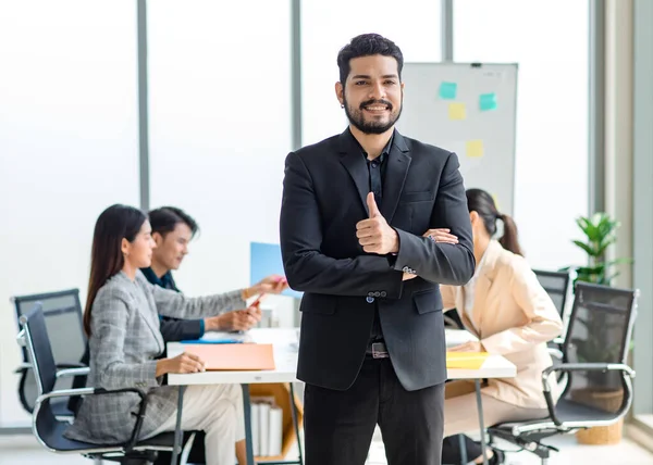 Portrait shot of millennial Indian Asian professional successful bearded businessman in formal suit standing posing smiling crossed arms in meeting room while employee and colleague brainstorming