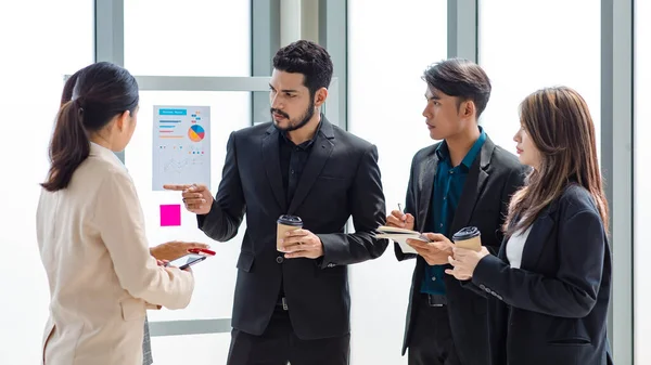 Group of millennial Asian Indian male female businessman businesswoman in formal suit standing holding tablet computer discussing sharing business ideas in multinational company office meeting room.