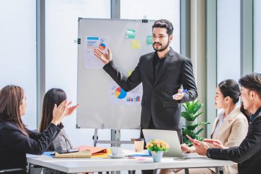 Businessman manager presenter in formal suit standing holding pen pointing at graph chart document on whiteboard presenting company information to Asian male female colleagues in meeting room.
