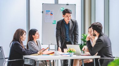 Businessman manager presenter in formal suit standing holding pen pointing at graph chart document on whiteboard presenting company information to Asian male female colleagues in meeting room.