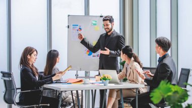 Indian businessman manager presenter in formal suit standing holding pen pointing at graph chart document on whiteboard presenting company information to Asian male female colleagues in meeting room.