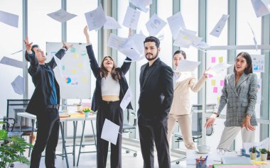 Portrait shot of millennial Indian Asian professional successful bearded businessman in formal suit standing posing while male female colleague celebrating throwing paperwork documents up in air.