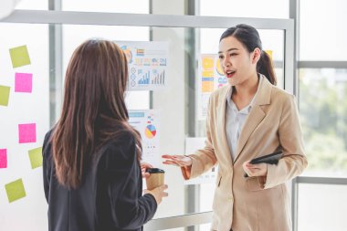 Two young millennial Asian female businesswomen in formal suit standing discussing talking sharing business ideas together in front glass board with graph chart information data paperwork documents.