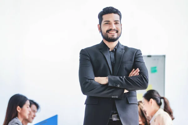 Portrait shot of millennial Indian Asian professional successful bearded businessman in formal suit standing posing smiling crossed arms in meeting room while employee and colleague brainstorming