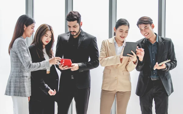 Group of millennial Asian Indian male female businessman businesswoman in formal suit standing holding tablet computer discussing sharing business ideas in multinational company office meeting room.