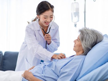 Closeup shot of unrecognizable unknown doctor in white lab coat with stethoscope hand holding comforting supporting old senior unhealthy patient in hospital uniform laying down on bed in ward room.
