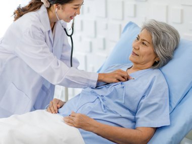 Closeup shot of unrecognizable unknown doctor in white lab coat with stethoscope hand holding comforting supporting old senior unhealthy patient in hospital uniform laying down on bed in ward room.