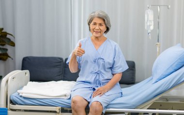 Portrait shot Asian old senior healthy gray hair female pensioner patient in blue hospital uniform sitting smiling holding hand showing thumb up on clinic bed in ward room with saline solution pole.