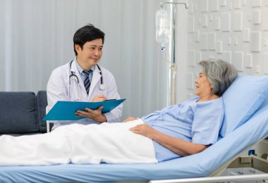 Asian male professional doctor practitioner in white lab coat and stethoscope writing notes when visiting monitoring old senior female pensioner grandma patient in hospital uniform laying down on bed.