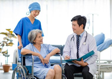Asian male professional doctor practitioner in white lab coat and stethoscope writing notes when visiting monitoring examining old senior female pensioner patient sitting on wheelchair in hospital.