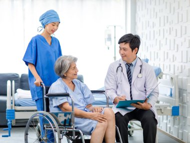 Asian male professional doctor practitioner in white lab coat and stethoscope writing notes when visiting monitoring examining old senior female pensioner patient sitting on wheelchair in hospital.