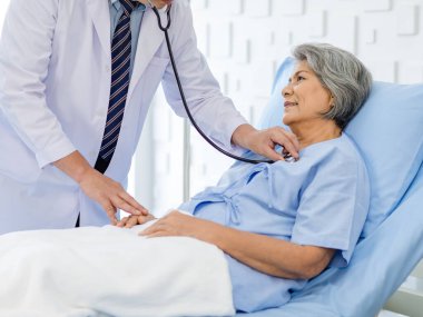 Asian male professional doctor practitioner in white lab coat using stethoscope listening examining heartbeat pulse of happy old senior female pensioner patient in hospital uniform lay down on bed.