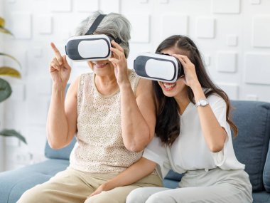 Asian young cheerful daughter wearing VR virtual reality goggles headset sitting smiling on cozy sofa in living room teaching old senior grey hair pensioner mother playing streaming 3d game online.