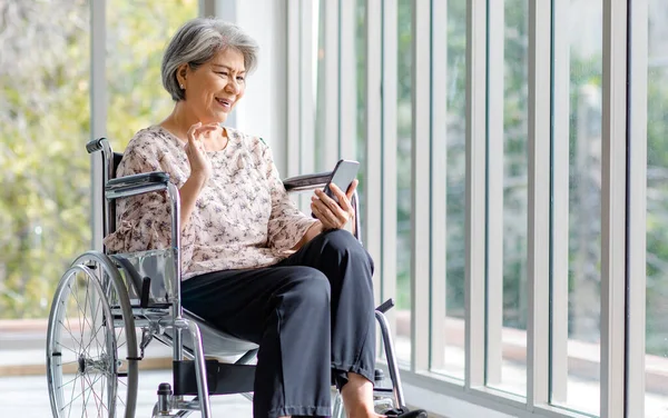 Asian happy cheerful old senior healthy gray hair retired pensioner disability handicapped grandmother sitting smiling on wheelchair using touchscreen smartphone surfing browsing social media online.