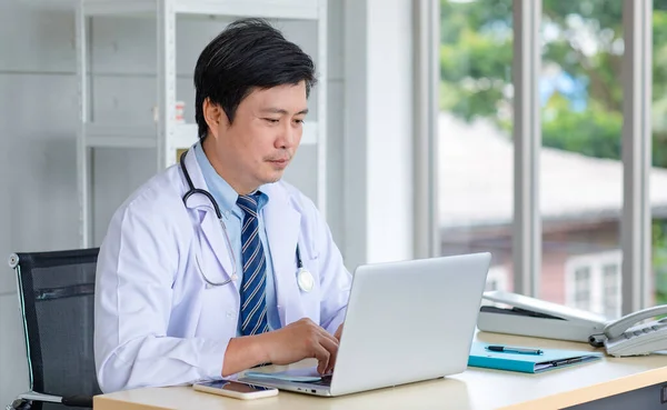 Asian middle aged male professional successful doctor practitioner in white lab coat with stethoscope sitting working at workstation desk typing laptop notebook computer in clinic hospital office.