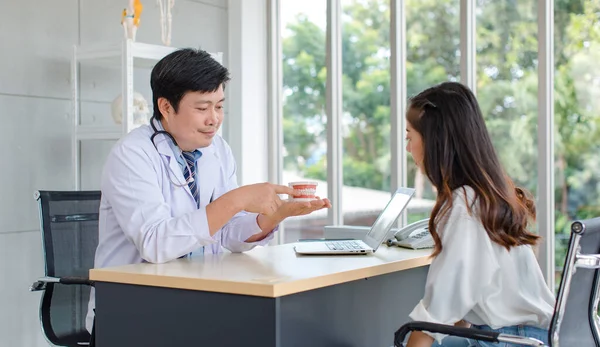 Asian middle aged male professional dentist in white lab coat with stethoscope sitting holding showing pointing denture model explaining to female patient at working desk in dental clinic office.