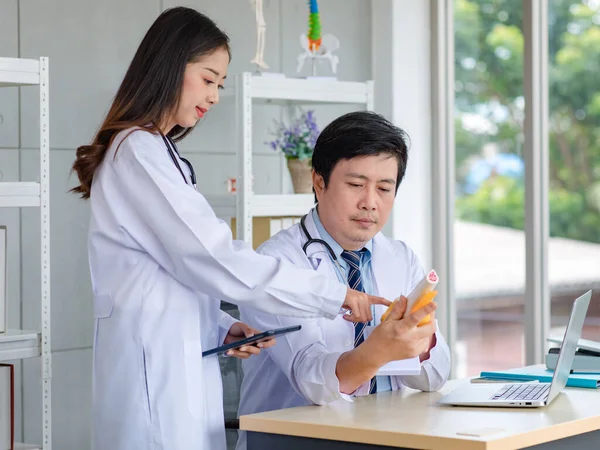 Asian young female doctor standing pointing skeleton model discussing with middle aged male professional colleague practitioner in white lab coat with stethoscope sitting at working desk in office.