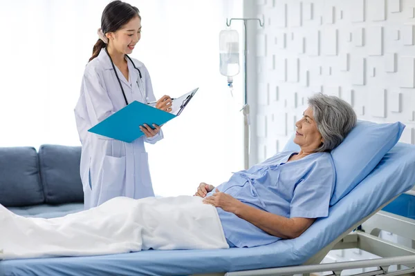 Asian female professional doctor practitioner in white lab coat with stethoscope standing smiling holding paperwork document folder visiting old senior patient in hospital uniform laying down on bed.