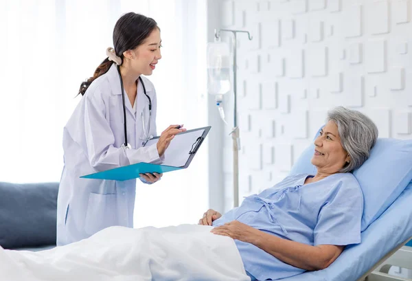 Asian female professional doctor practitioner in white lab coat with stethoscope standing smiling holding paperwork document folder visiting old senior patient in hospital uniform laying down on bed.