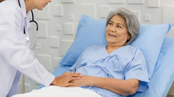 Closeup shot of unrecognizable unknown doctor in white lab coat with stethoscope hand holding comforting supporting old senior unhealthy patient in hospital uniform laying down on bed in ward room.