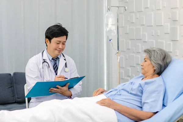 Asian male professional doctor practitioner in white lab coat and stethoscope writing notes when visiting monitoring old senior female pensioner grandma patient in hospital uniform laying down on bed.
