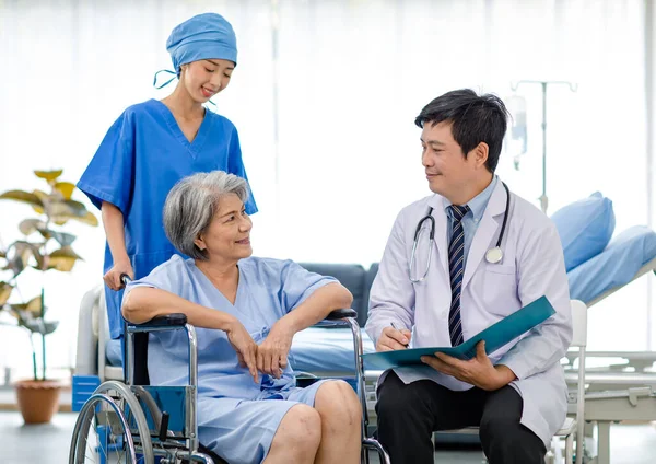 Asian male professional doctor practitioner in white lab coat and stethoscope writing notes when visiting monitoring examining old senior female pensioner patient sitting on wheelchair in hospital.
