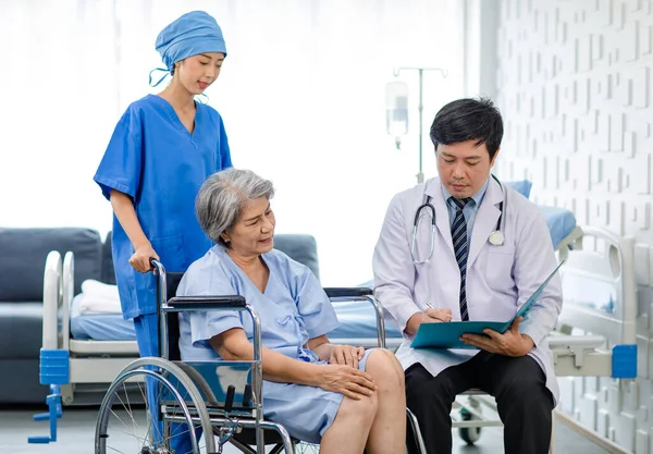 Asian male professional doctor practitioner in white lab coat and stethoscope writing notes when visiting monitoring examining old senior female pensioner patient sitting on wheelchair in hospital.