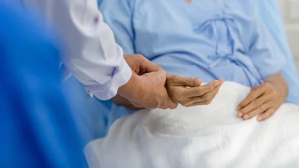Closeup shot hands of unrecognizable unknown doctor practitioner in white lab coat holding checking examining heartbeat pulse from patient in blue hospital uniform laying down on bed in ward room.