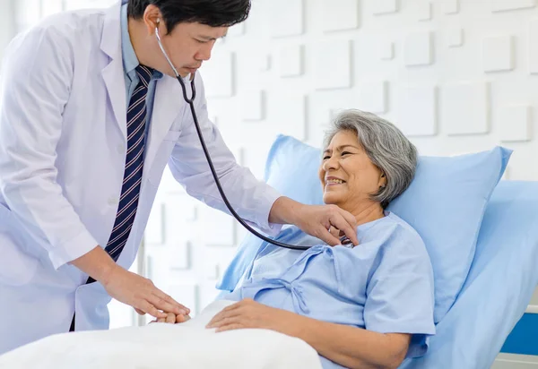Asian male professional doctor practitioner in white lab coat using stethoscope listening examining heartbeat pulse of happy old senior female pensioner patient in hospital uniform lay down on bed.